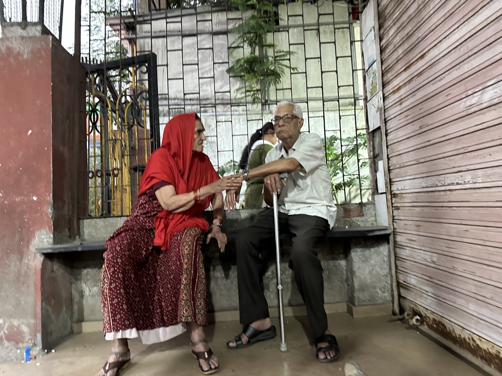 An elderly couple in conversation on a bench by a shuttered shopfront